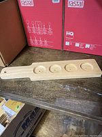 Single wooden beer flight paddle with four cup indentations and hole in handle, placed on wooden surface with boxes in background