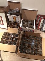 Five open cardboard boxes containing assorted beverage glasses including tall beer glasses, stemmed glasses, and short tumblers with textured bases. Glasses are organized in cardboard dividers for protection.