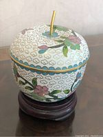 Front and side view of cloisonne lidded trinket box on wooden stand, showing apple shape with floral enamel decoration and gold stem handle