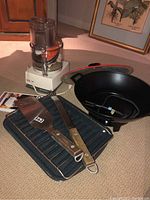 Full lot with food processor in background, wok with lid, broiler pan and utensils on carpeted floor.