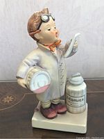 Front side view of Hummel Little Pharmacist figurine showing child holding a prescription and a bottle labeled 'Vitamins'.