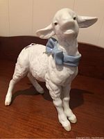 Front and side view of glazed white porcelain baby lamb figurine with a blue bow around the neck, standing on a wooden surface.