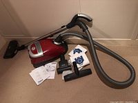 Red Miele canister vacuum with hose, telescopic wand, multiple attachments, vacuum bags and manuals arranged on carpet