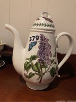 Full view of white ceramic coffee pot decorated with lilac flower and butterfly botanical illustrations on side with spout and handle visible, marked '379'.
