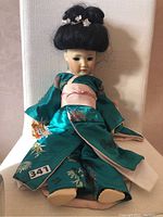 Full view of the doll seated, dressed in teal silk kimono with pink sash, porcelain head, and dark hair styled up.