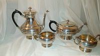 Four-piece silver plated Art Deco tea and coffee set shown together against a white fabric background. Includes coffee pot, teapot, creamer, and sugar bowl with black handles on pots.
