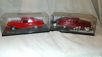 Two model cars in clear plastic display cases. One is rectangular with a shiny red '64 Ford Fairlane model and a small red gas pump accessory. The other case contains a classic deep red model car with an exposed engine model and several trophies.