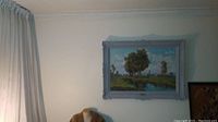 Full view of framed landscape painting on wall showing river, trees, meadow and homes.