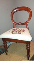 Side angle of antique balloon back chair showing polished wooden frame and needlepoint floral embroidered seat.