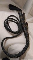 Black studded leather whip coiled on white surface showing braided leather and studded handle with loop handles for hanging or grip.