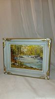 Frontal photo showing entire framed oil painting of a river and forest landscape with light blue and gold cornered frame.