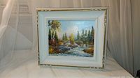 Framed oil painting showing full artwork of Canadian forest landscape with river and rocks inside a white and gold frame.