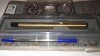 Front view of unopened original packaging showing gold pen, keychain, and two ink refills with blue caps in clear plastic box.