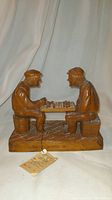Wood carving sculpture of two men playing checkers, viewed frontally.