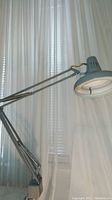 Side view of the drafting lamp showing the large round gray lamp head and double arm with springs for tension and swivel attached to base clamp.