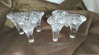 Pair of clear textured glass candleholders with three legs and three candle sockets, shown on brown fabric background.