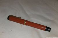 Full view of capped pen lying on fabric surface showing orange body and black ends with gold clip and band.
