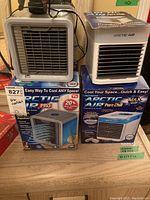 Two Arctic Air coolers side by side with their original boxes, placed on a wooden floor: Arctic Air Pro and Arctic Air Pure Chill.