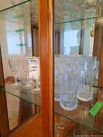 Crystal stemmed wine glasses and tall soft drink glasses arranged on glass shelves inside a wooden cabinet, visible chips on some glasses.