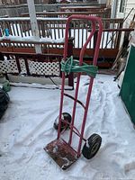 Metal dolly shown standing on snow-covered surface with green rope tied around handle, showing front side and wheels.