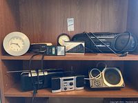 Wide shot of seven clocks, clock radios, and a boombox arranged on wooden shelves.