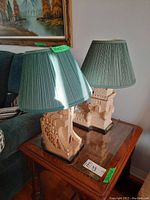 Two decorative table lamps with castle turret bases and green pleated fabric shades on wooden side table next to sofa.