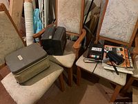 Photo showing vintage Sony reel to reel tape recorder in beige carrying case with cables and accessories, Bell & Howell movie camera, and projector on chairs