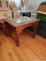Wood side table shown with clear glass top, placed on hardwood floor next to a green couch with cardboard boxes in background.