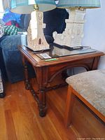 Full view of wooden side table with glass top, showing turned legs and arched support design, situated next to chair and sofa.