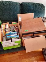 Two cardboard boxes filled with assorted VHS tapes and two wood-finish cases for tape storage on a green sofa and wooden floor.