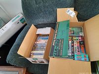 Two boxes filled with multiple VHS tapes stacked inside original and cardboard cases on a couch.