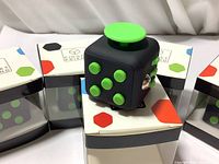 Black and green fidget cube on top of clear retail packaging boxes, showing one side with six green buttons and one large green circular button on top.