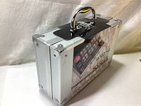 Closed silver metal train case with black handle and latch, product packaging visible on front.