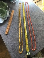 Two long beaded necklaces, one yellow and one orange, placed on a grey countertop with rulers for scale.
