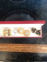 Three pairs of clip-on earrings arranged in a white cushioned box with red edges, shown alongside ruler for size context. Earrings wrapped with cloth bandage to reduce metal allergy. First pair butterfly style with rhinestones, second pair sunburst with rhinestone center, third pair black and gold twisted knot.