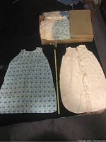 Photo showing two baby sleep sacks — blue patterned and white ruffled, plus a box of folded baby clothes.