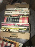 Photo of about 15 Better Homes & Gardens cookbooks stacked inside a cardboard box showing various titles clearly.