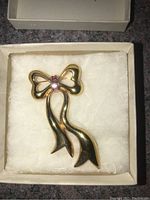 Gold-tone bow-shaped pin with ruby-colored crystals displayed in a white jewelry box on a cushioned pad.