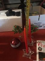 Photo showing two red taper candles in star-shaped glass holders, a round red ornament, two crystal glasses with holly decoration, and visible holiday baking cookbooks in background.