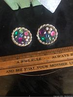 Photo of two round clip-on earrings showing multicolor rhinestones and imitation pearls with twisted gold-tone rope border on a black background; a wooden ruler is below for scale.