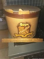 Vintage Biltmore hat box with visible tape repair on lid and logo on front, ruler for scale showing about 10 inch height and 13 inch diameter