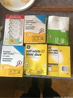 Six boxes of light bulbs on a table including Sunbeam, Signal, and GE brands with 60 watt soft white and frosted bulbs