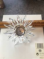 Front view of Swarovski crystal sunburst clock lying on a white surface with a ruler for size reference. Shows clear crystal rays and clock face.
