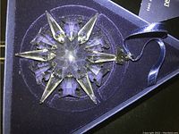 Close-up photo of the clear crystal snowflake ornament in its blue velvet-lined triangular box showing detailed facets and attached blue ribbon.