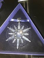 Clear crystal snowflake ornament with multiple pointed arms inside a blue triangular box with fitted foam insert.