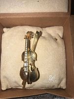 Front view of gold-tone violin pin brooch with clear Swarovski crystals on fretboard, resting on a beige cushion inside a box with Swarovski logo.