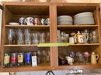 Wooden cabinet shelves holding the assortment of mugs, glasses, plates, teapot, and bottles.