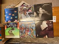 Top-down view of 7 assorted vinyl records featuring covers with artists like Stevie Wonder, Elton John, cartoon hockey players, John Lennon & Yoko Ono, and others.