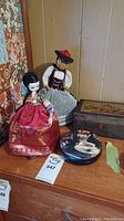 Photo showing two dolls in traditional costumes, one wooden box and one round tin box on a wooden surface with wallpaper background