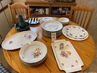 Wide shot of assorted porcelain plates and serving trays on wooden table showing floral patterns and serving pieces.